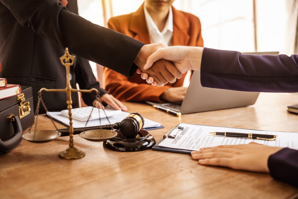 A lawyer in the background overseeing two people shaking hands as part of a plea deal agreement