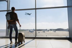 Young man is standing near window at the airport and watching plane before departure.